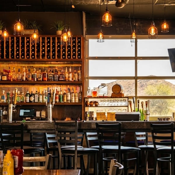 A well-stocked bar with bottles, glasses, and a person sitting at the counter, with mountains visible outside.
