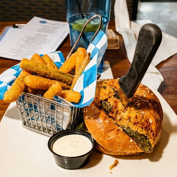 A burger with a knife, a basket of fried pickles, and dipping sauce on a wooden table.
