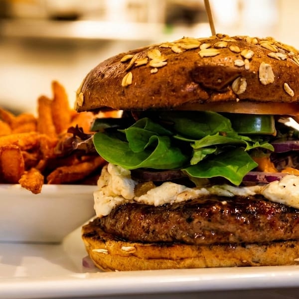 A gourmet burger with an oat-topped bun, lettuce, onions, and sauce, served with sweet potato fries.