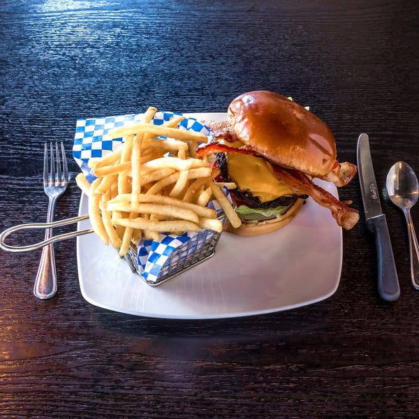 Bacon cheeseburger and fries in a basket on a white plate with cutlery on a dark table.